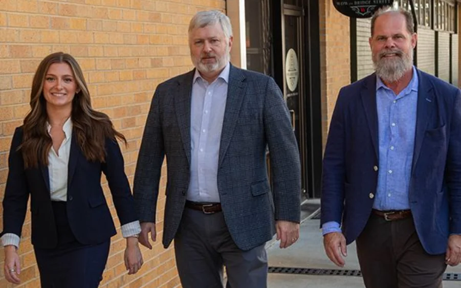 Three people walking: woman in navy suit, two men in jackets; outdoors, brick wall.